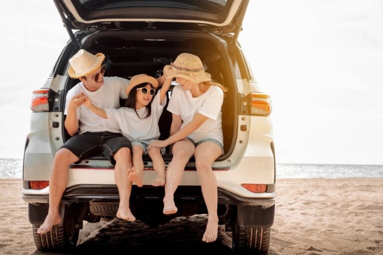 A happy family wearing hats and sunglasses, sitting in the open trunk of a car, enjoying a sunny day at the beach