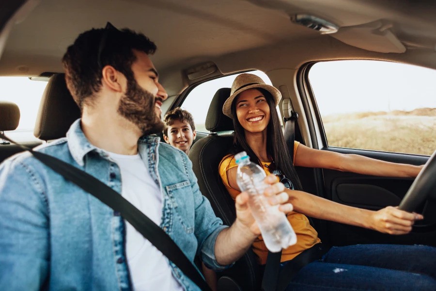 A happy family on a road trip, with a woman driving, a man holding a water bottle in the passenger seat, and a smiling child sitting in the backseat.