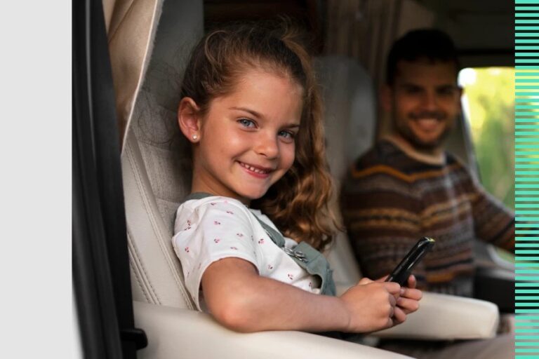 A smiling young girl sitting in a car seat holding a phone, with a man in the driver’s seat in the background.