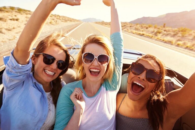 Three joyful women wearing sunglasses, laughing and raising their arms while enjoying a road trip in a convertible under the bright sun.