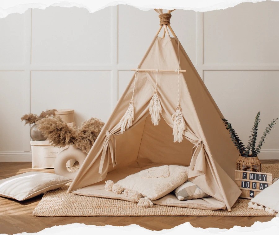 A beige kids' teepee tent decorated with macramé accents, surrounded by cozy cushions, pampas grass, and a woven basket in a stylish, minimalist playroom.